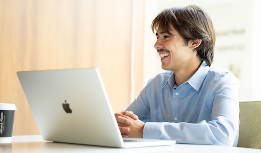 Student sitting at a desk, smiling to someone across the room with his laptop open